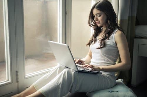 Woman using her laptop while relaxing beside a windowsill